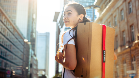 Anerkennung Kategorie Unternehmen 2024 Fotografie einer jungen Frau, die im städtischen Raum mithilfe des roten Gurtsystems einen mittelgroßen Paketkarton auf dem Rücken trägt.